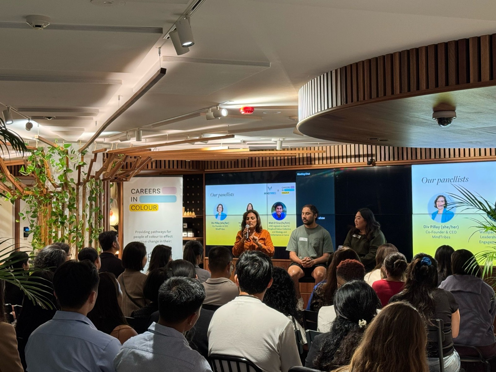 A panel discussion hosted by Careers in Colour, featuring speakers seated on stage before an audience. Event banners and presentation screens display panelist information in a modern, well-lit venue with greenery and wooden accents.